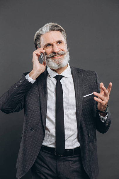 Liiketoiminta ja muodikas parrakas kypsä mies harmaassa puvussa puhuu puhelimitse ja hymyilee harmaalla taustalla. - Valokuva, kuva