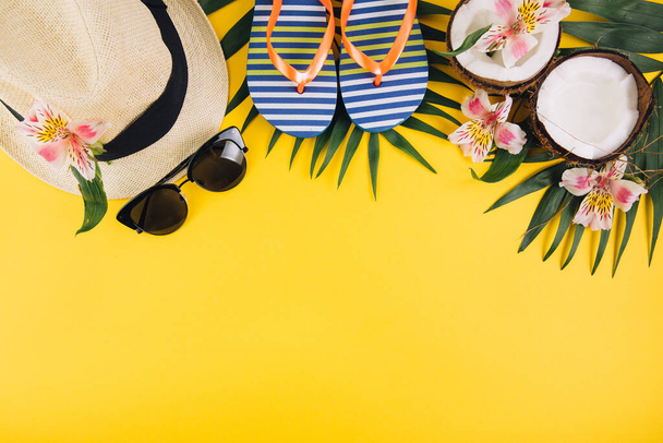 熱帯ヤシの葉、帽子、スリッパ、サングラス、黄色の背景にココナッツ。夏のコンセプト。フラットレイアウト、トップビュー、コピースペース - 写真・画像