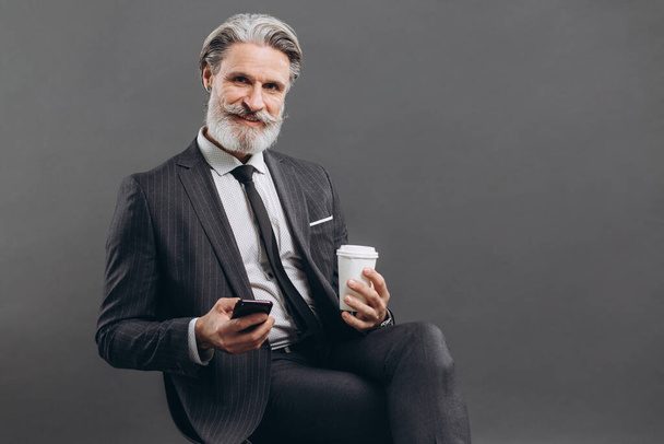 Бізнес і модний бородатий зрілий чоловік у сірому костюмі, що сидить і використовує смартфон на сірому фоні
. - Фото, зображення