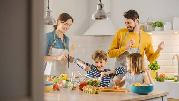 In Kitchen: Family of Four Cooking Together Healthy Dinner, Fool Around and Dance. Mother, Father, Little Boy and Girl, Preparing Salads, Cutting Vegetables. Cute Children Helping their Caring Parents - Foto, imagen