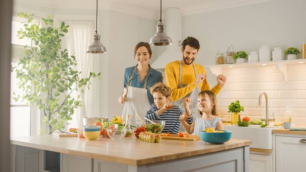 In Kitchen: Family of Four Cooking Together Healthy Dinner, Fool Around and Dance. Mother, Father, Little Boy and Girl, Preparing Salads, Cutting Vegetables. Cute Children Helping their Caring Parents - Фото, изображение