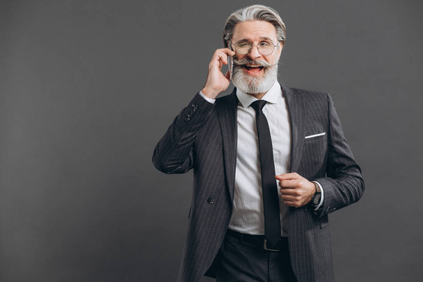 Business and fashionable bearded mature man in a gray suit smiling and talking by phone on the grey background. - Foto, Imagen