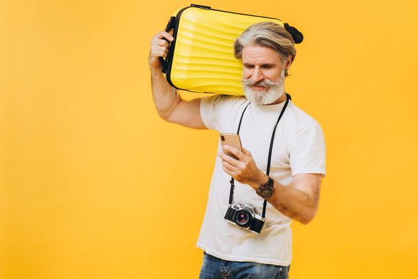 Stilvoller bärtiger Mann, der lacht, einen gelben Koffer in der Hand hält und auf das Telefon schaut, das sich ein Reiseland aussucht. - Foto, Bild