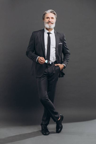 Negócios e homem maduro barbudo na moda em um terno cinza segurando seus óculos e sorrindo em um fundo cinza
. - Foto, Imagem