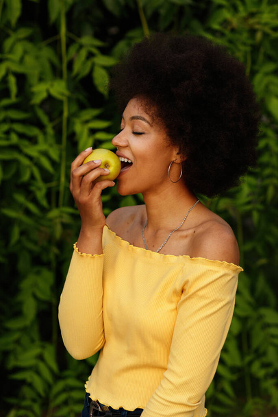 暗い肌の少女は緑の背景に緑のリンゴを食べます。夏 - 写真・画像
