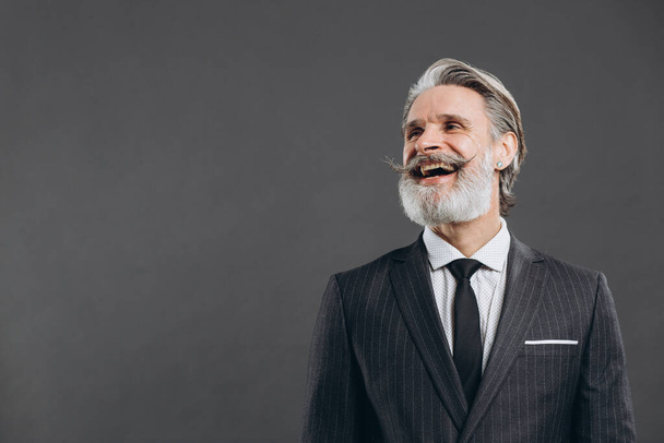 Porträt eines lächelnden und modisch reifen bärtigen Mannes im grauen Anzug. Business- und trendiges Nahaufnahmekonzept auf grauem Hintergrund. - Foto, Bild