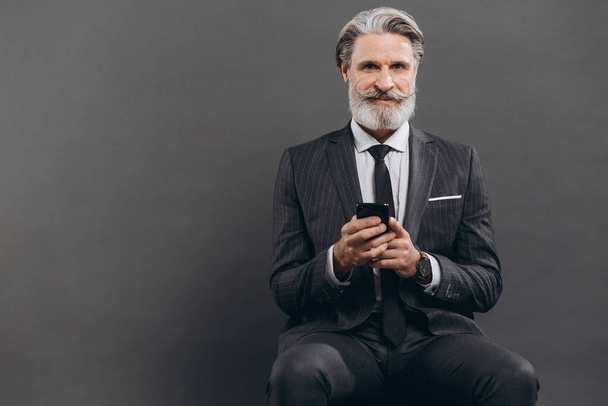 Бізнес і модний бородатий зрілий чоловік у сірому костюмі, що сидить і використовує смартфон на сірому фоні
. - Фото, зображення