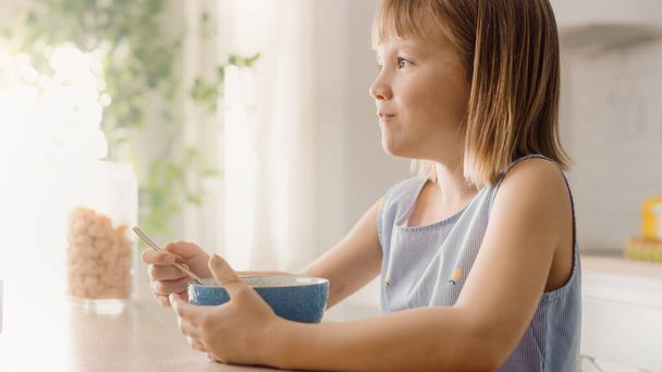 Breakfast in the Kitchen: Portrait of Adorable Little Girl Eating Healthy Granola Cereal with Milk out of Bowl. Cute Little Girl having Breakfast. In the Background Caring Mother Prepares Food - Valokuva, kuva