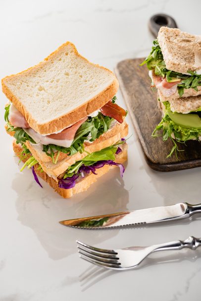 Свежие сэндвичи с мясом на мраморной белой поверхности со столовыми приборами и деревянной доской
 - Фото, изображение