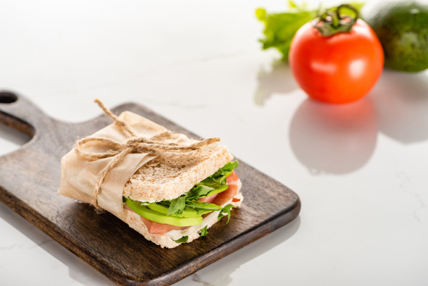selective focus of fresh green sandwich with avocado and prosciutto on wooden cutting board on white marble surface - Foto, immagini