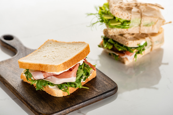selective focus of fresh green sandwich with prosciutto on wooden cutting board on white marble surface - 写真・画像