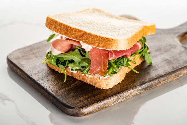 selective focus of fresh green sandwich with arugula and prosciutto on wooden cutting board on white marble surface - Fotó, kép