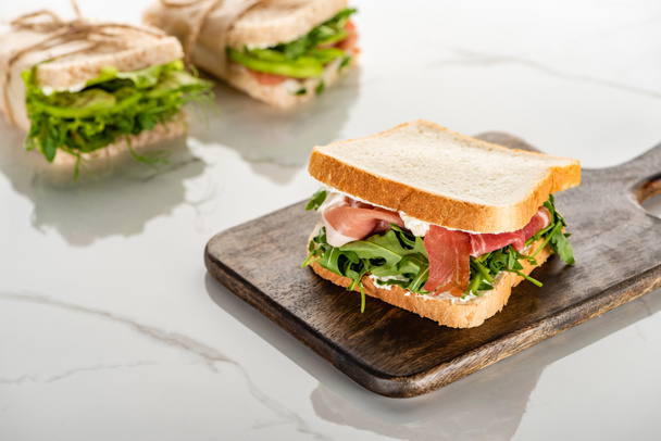 selective focus of fresh green sandwich with arugula and prosciutto on wooden cutting board on white marble surface - Foto, immagini