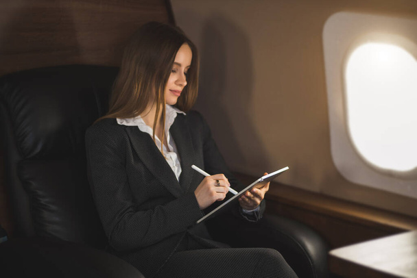 Nice young businesswoman in formal attire, with makeup, looking at screen of tablet with satisfied expression, working remotely while flying on private jet. Business, traveling and technology. - Фото, зображення