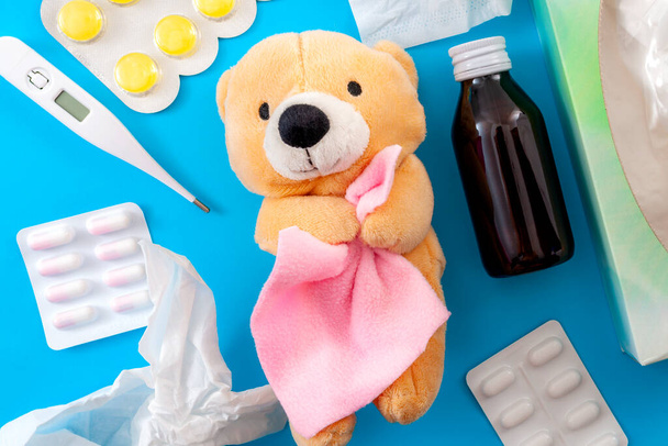 Cold and flu treatment and pediatrics medicine concept with a teddy bear holding a blanket surrounded by  a thermometer, bottle of cough syrup, sore throat lozenges, pills, capsules and paper tissue - Photo, Image