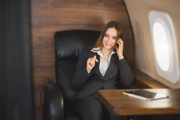 Cheerful happy brown haired business lady in white blouse and formal suit, sitting in comfortable seat, laughing talking on mobile phone during flight on first class plane. - Fotografie, Obrázek