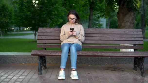 Een jonge vrouw zit op een bank met een telefoon in haar handen. Het meisje in de ochtend in het park is in correspondentie per telefoon. - Video