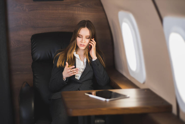 Attractive nervous young caucasian businesswoman is talking on the phone with an irritated facial expression, gesturing, sitting on a private plane. Business, emotions concept. - Photo, Image