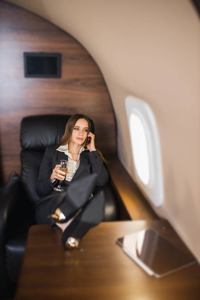 Confident self assured young businesswoman sitting in private plane with her feet on table, talking on mobile phone, holding glass of champagne, wearing formal black suit, high heel shoes. - Foto, afbeelding
