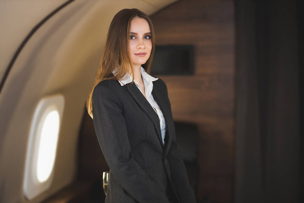 Beautiful caucasian young brown haired lady with nice makeup, wearing stylish black suit, looks down, touches her hair and gently smiles, poses in private aircraft. Traveling at first class. - Zdjęcie, obraz