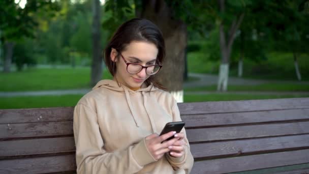 Μια νεαρή γυναίκα κάθεται σε ένα παγκάκι με ένα τηλέφωνο στα χέρια της. Το κορίτσι το πρωί στο πάρκο είναι σε αλληλογραφία μέσω τηλεφώνου closeup. - Πλάνα, βίντεο