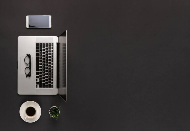 オープンノートパソコン、コーヒーカップ、コピースペースの抽象的な暗い黒い机の背景にスマートフォンのトップビュー。ミニマルモダンオフィスのコンセプト. - 写真・画像