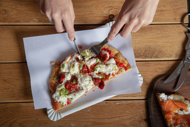 Las manos de una mujer cortaron un pedazo de pizza en una mesa de madera. La pizza italiana se come con un tenedor y un cuchillo. Primer plano
 - Foto, Imagen