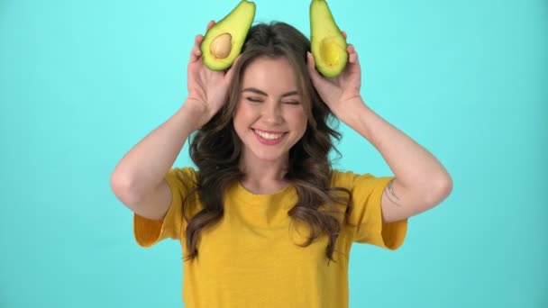 A funny young woman wearing a yellow t-shirt is having fun posing with avocado standing isolated over blue background - Filmati, video
