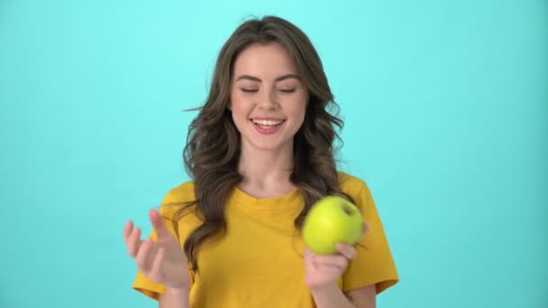 A pretty joyful young woman wearing a yellow t-shirt is posing with an apple standing isolated over blue background - Video