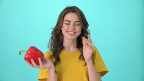 A smiling young woman wearing a yellow t-shirt is playing with red pepper standing isolated over blue background - Metraje, vídeo