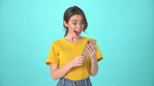 A surprised young woman wearing yellow t-shirt is looking to her smartphone while doing a winner gesture standing isolated over blue background - Séquence, vidéo