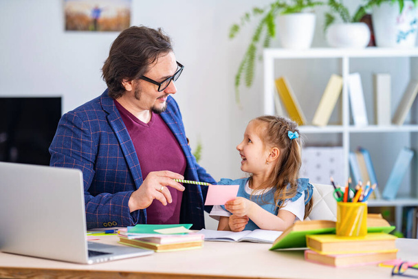 cute little girl is engaged in lessons with a teacher at home. Primary school tutor. Preschool education. Dad helps with homework. Individual training. - Foto, afbeelding