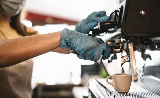 Young woman side view making coffee espresso while wearing surgical mask and gloves for preventing corona virus spread - Bar owner safety working - Hot beverages and covid-19 rules concept - Photo, Image
