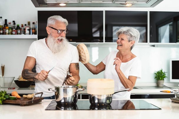 Happy senior couple having fun cooking together at home - Elderly people preparing health lunch in modern kitchen - Retired lifestyle family time and food nutrition concept - Foto, imagen