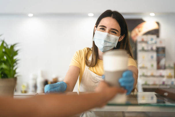 Bar owner working only with take away orders during corona virus outbreak - Young woman worker wearing face surgical mask giving coffee to customer - Healthcare and drinks concept - Photo, image