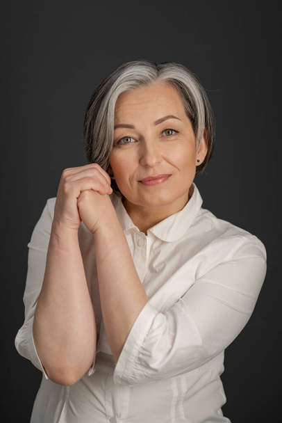 Aged woman charmingly smiles tilting head slightly and clasped hands near face. Beautiful lady in white shirt Isolated on gray - Valokuva, kuva