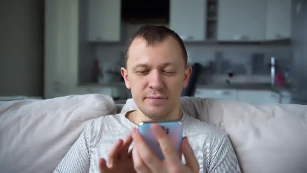 Homme souriant assis sur le canapé avec un téléphone portable dans ses mains, met likes, communication sur Internet - Séquence, vidéo