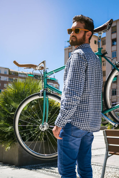 Hombre guapo con gafas de sol sosteniendo una bicicleta en su hombro en la calle. Fotografía horizontal al aire libre, estilo hipster con camisa a cuadros
 - Foto, imagen
