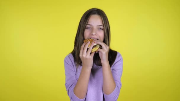 Tiener meisje in een roze trui eet een hamburger op gele achtergrond met kopieerruimte - Video