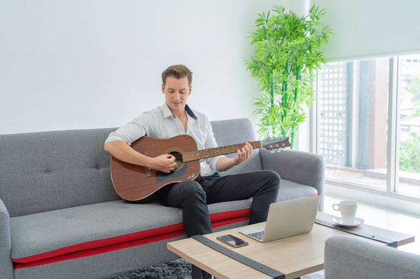 Biznes blondynka, biały muzyk, biały mężczyzna studiuje, ćwiczy i uczy się grać na gitarze w domu z laptopa notebooka komputerowego w Internecie w gitarzysta oglądając samouczek online. Muzyka na żywo. - Zdjęcie, obraz