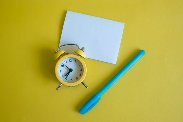 yellow alarm clock, blank white sheet and blue pen on yellow background top view - Photo, Image