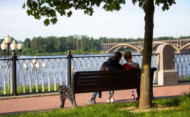 Una joven pareja está sentada en un banco de madera bajo un árbol cerca del río
. - Foto, Imagen