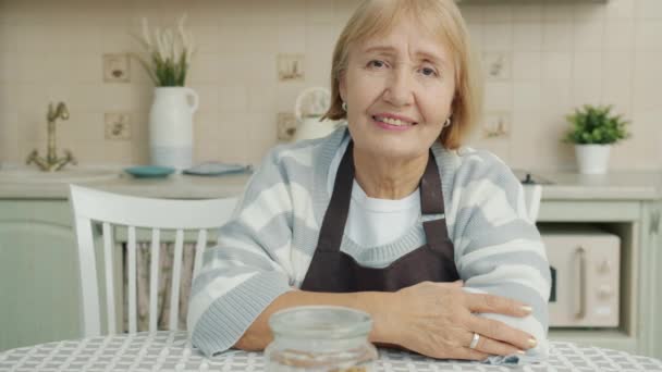 Portrait of beautiful elderly woman wearing apron sitting in kitchen alone and looking at camera - Imágenes, Vídeo