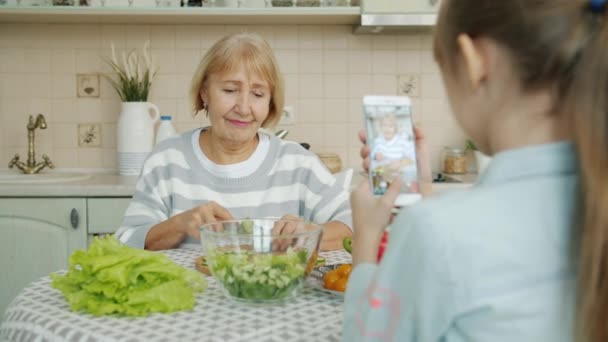 Little girl taking pictures of granny cooking salad then posing for camera with cucumber slices - Séquence, vidéo