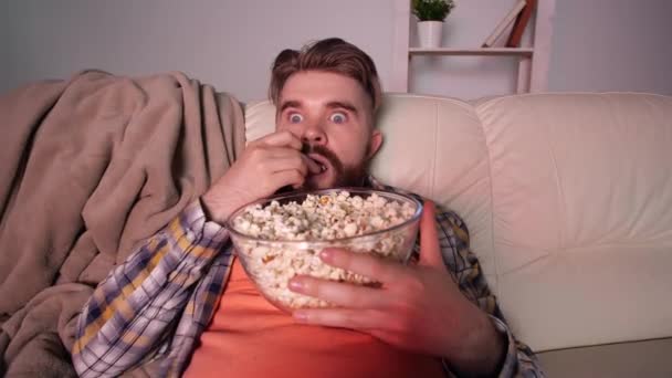 Ev sinema konsepti. Şok olmuş ve korkmuş bir adam televizyon seyrediyor ve patlamış mısır yiyor. - Video, Çekim