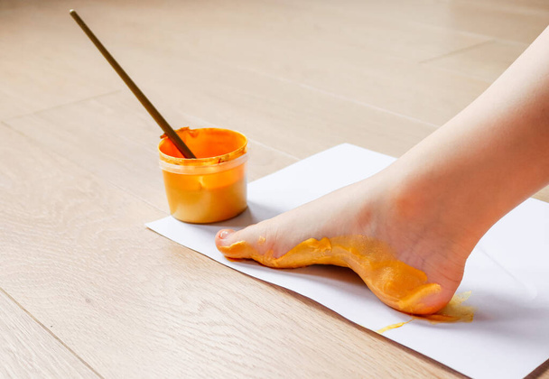 vue latérale d'une jambe d'enfant sur une feuille de papier blanche avec un pot de peinture jaune et un pinceau dedans. - Photo, image