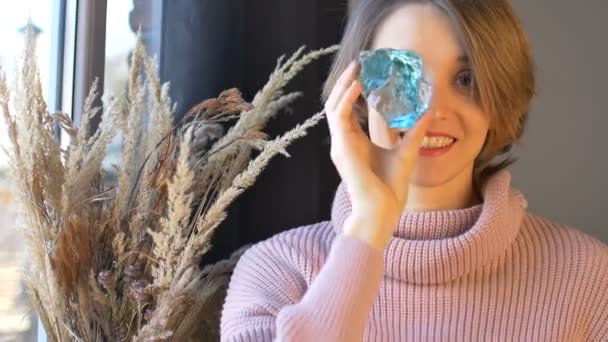 Nuori nainen, jolla on lyhyet vaaleat hiukset, pitää kädessään läpinäkyvää sinistä lasia seisomassa ikkunan lähellä spikelets-maljakon kanssa. Naisten terveyskäsite, taide - Materiaali, video