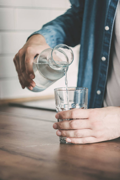 The man pours himself a glass of clean water. A carafe of water on the kitchen wooden table. Bottle of water. the concept of a healthy lifestyle - Valokuva, kuva