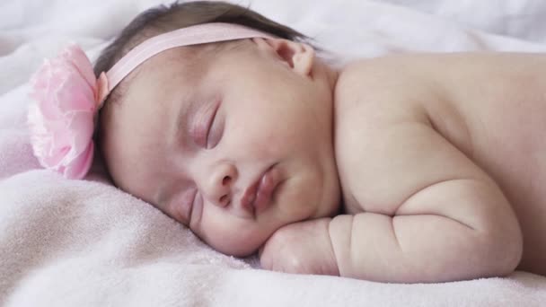 βρεφική ηλικία, παιδική ηλικία, ανάπτυξη, ιατρική και υγειονομική έννοια - γκρο πλαν πρόσωπο ενός νεογέννητου γυμνού κοριτσιού που κοιμάται ξαπλωμένη στο στομάχι της με έναν επίδεσμο και ένα λουλούδι στο κεφάλι της σε ροζ φόντο. - Πλάνα, βίντεο
