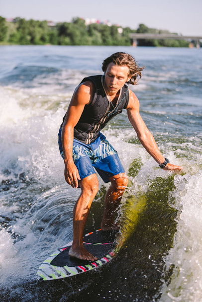Beautiful long haired blond wakesurf in vest on board along waves of lake. Athletic male athlete wakes surfing in summer on river. Wakesurf water sport. Surfer on wave. Man surfing on the Surfboard. - Valokuva, kuva